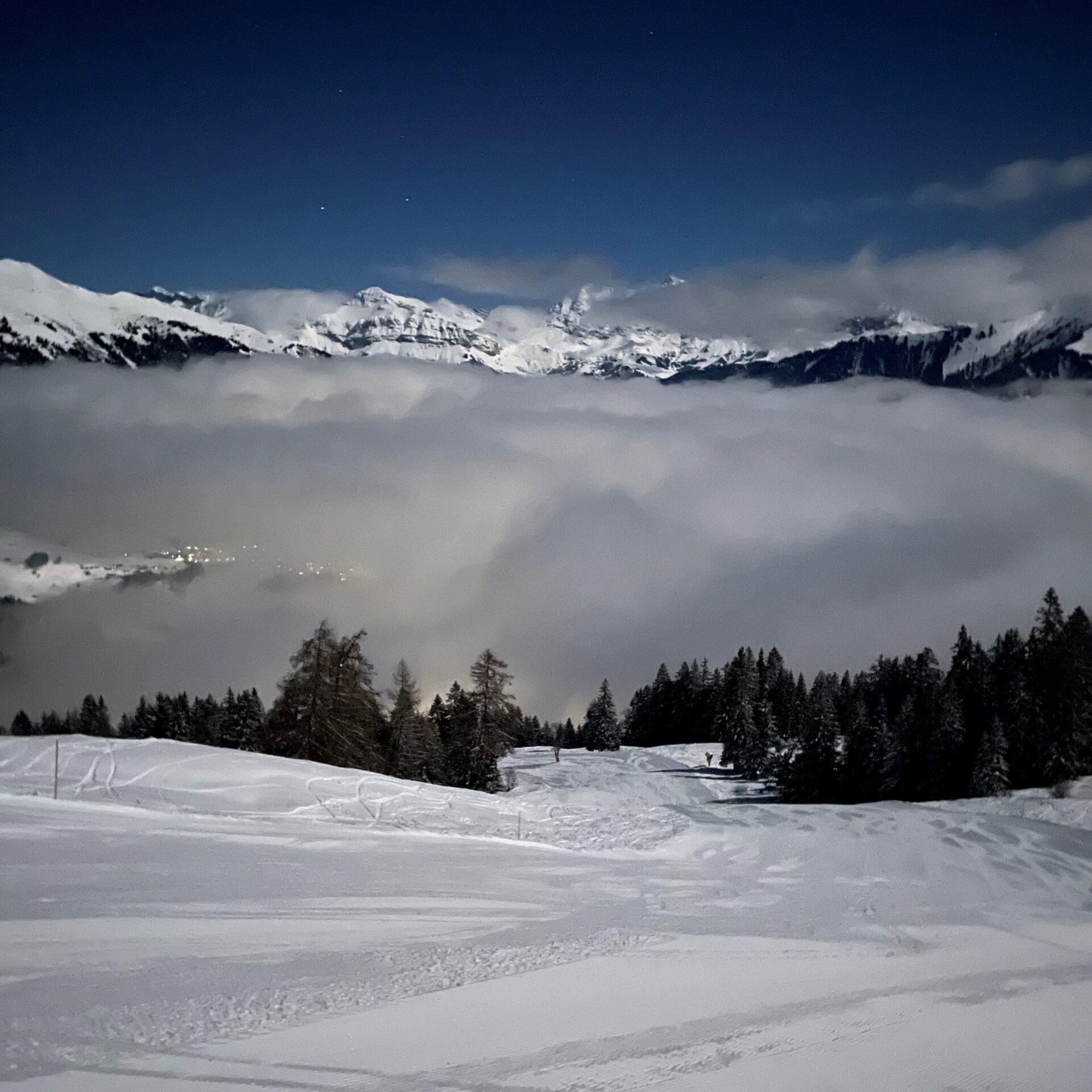 Blick von der Piste auf das Nebelmeer und die leuchtenden Berge bei der Vollmondnacht am Turneri