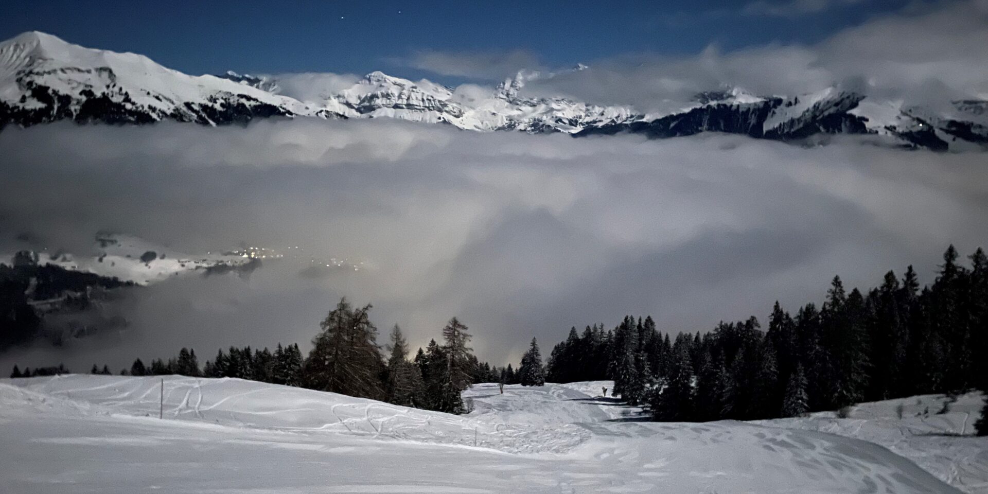 Blick von der Piste auf das Nebelmeer und die leuchtenden Berge bei der Vollmondnacht am Turneri