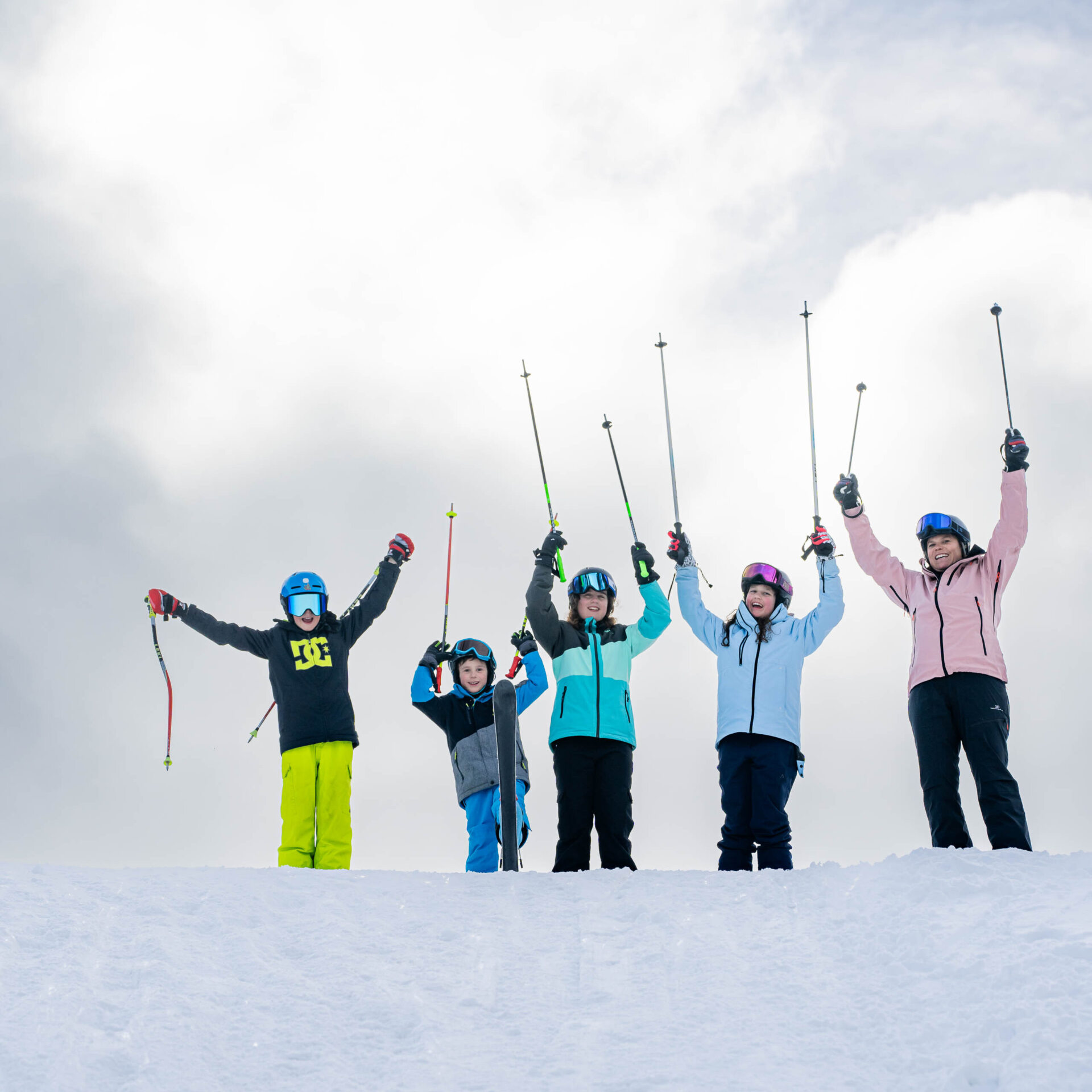 Eine Erwachsene und vier Kinder stehen nebeneinander und strecken ihre Skistoecke in die Luft