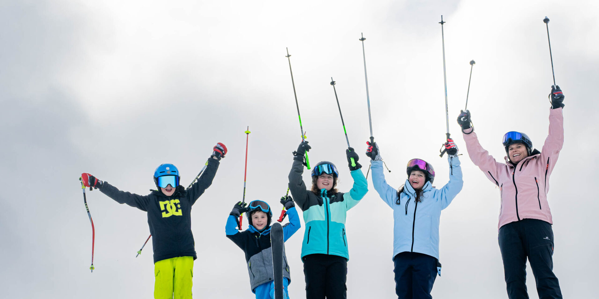 Eine Erwachsene und vier Kinder stehen nebeneinander und strecken ihre Skistoecke in die Luft