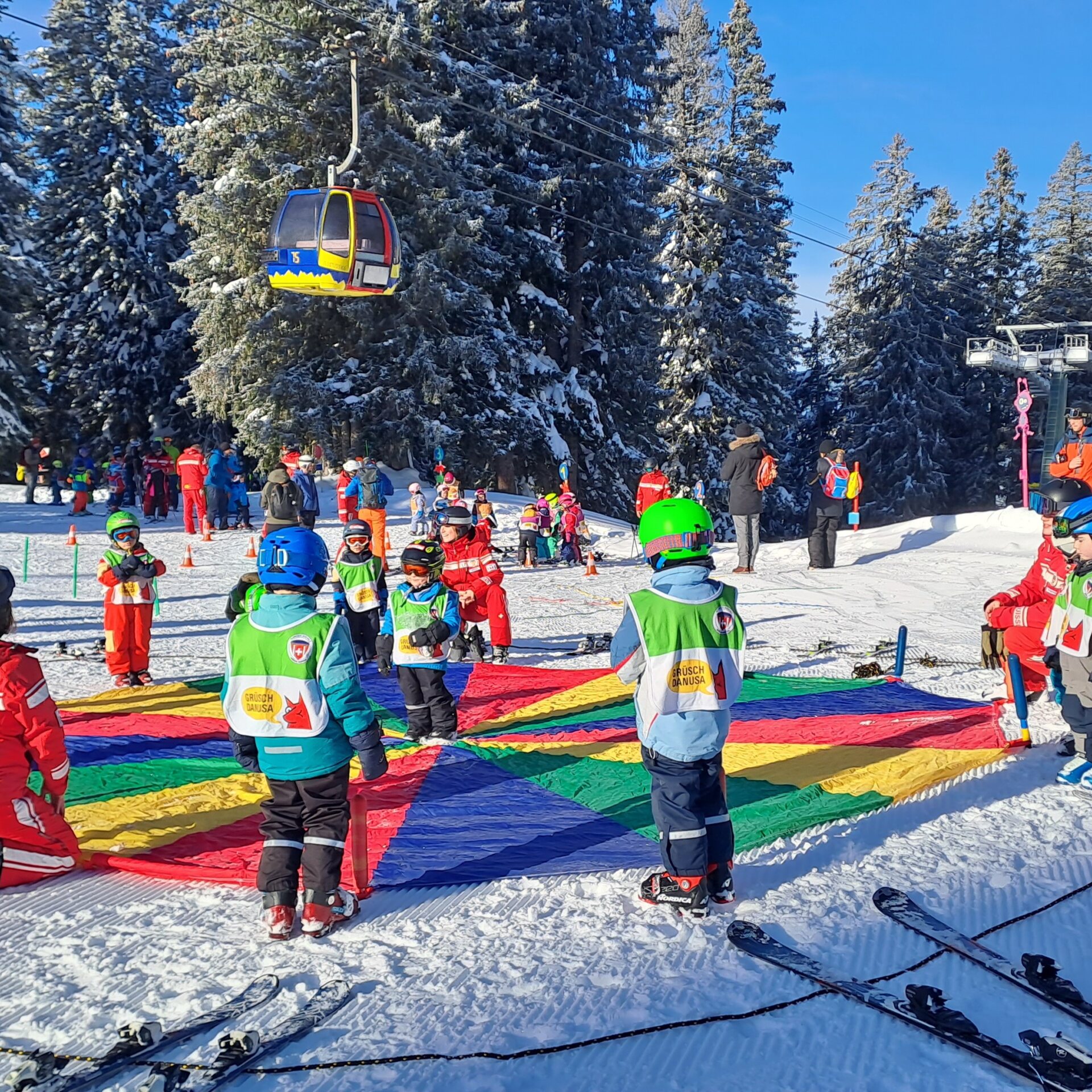 Skilehrerinnen und kleine Kinder stehen um ein Regenbogentuch und machen ein Spiel