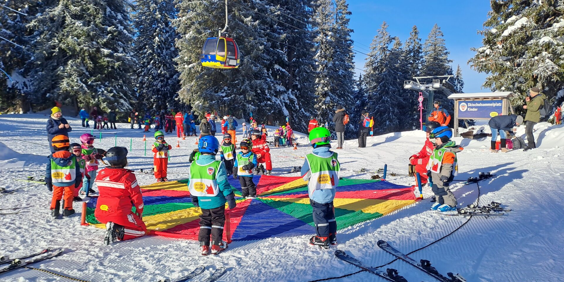 Skilehrerinnen und kleine Kinder stehen um ein Regenbogentuch und machen ein Spiel