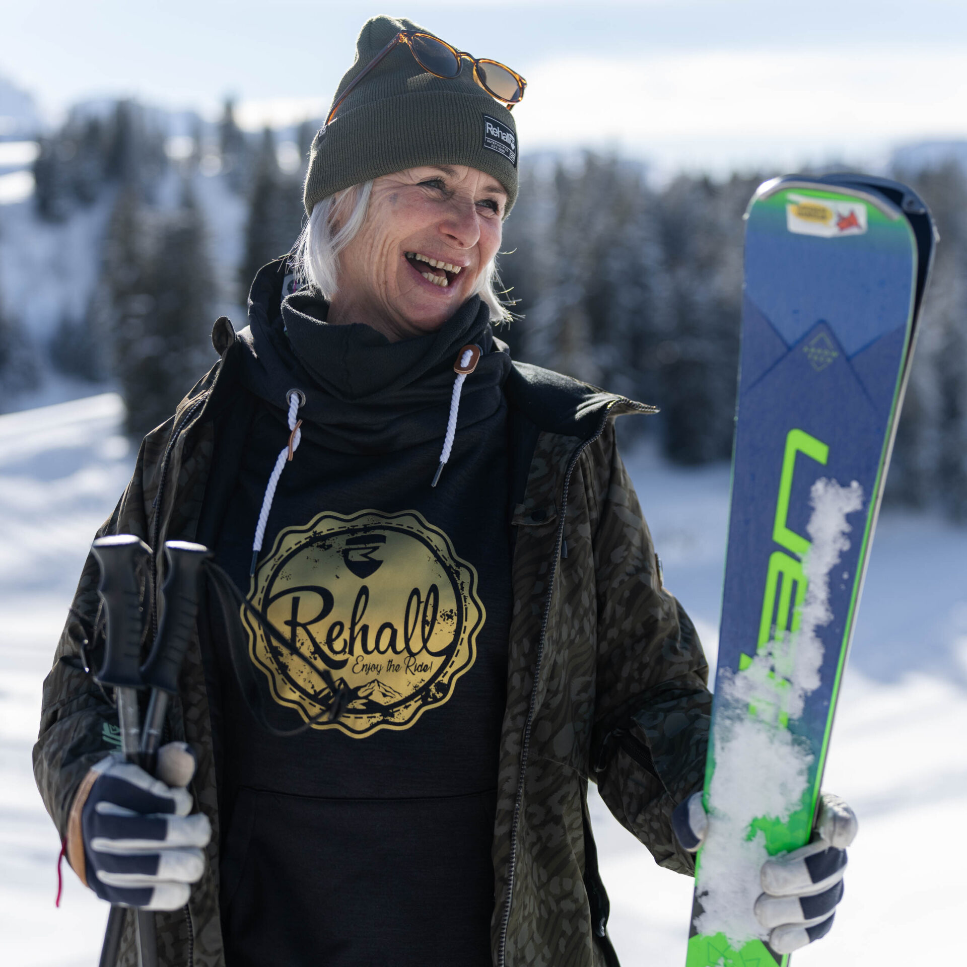 Skifahrerin steht mit offener Jacke in je einer Hand Ski und Stoecke schaut auf dei Seite und lacht