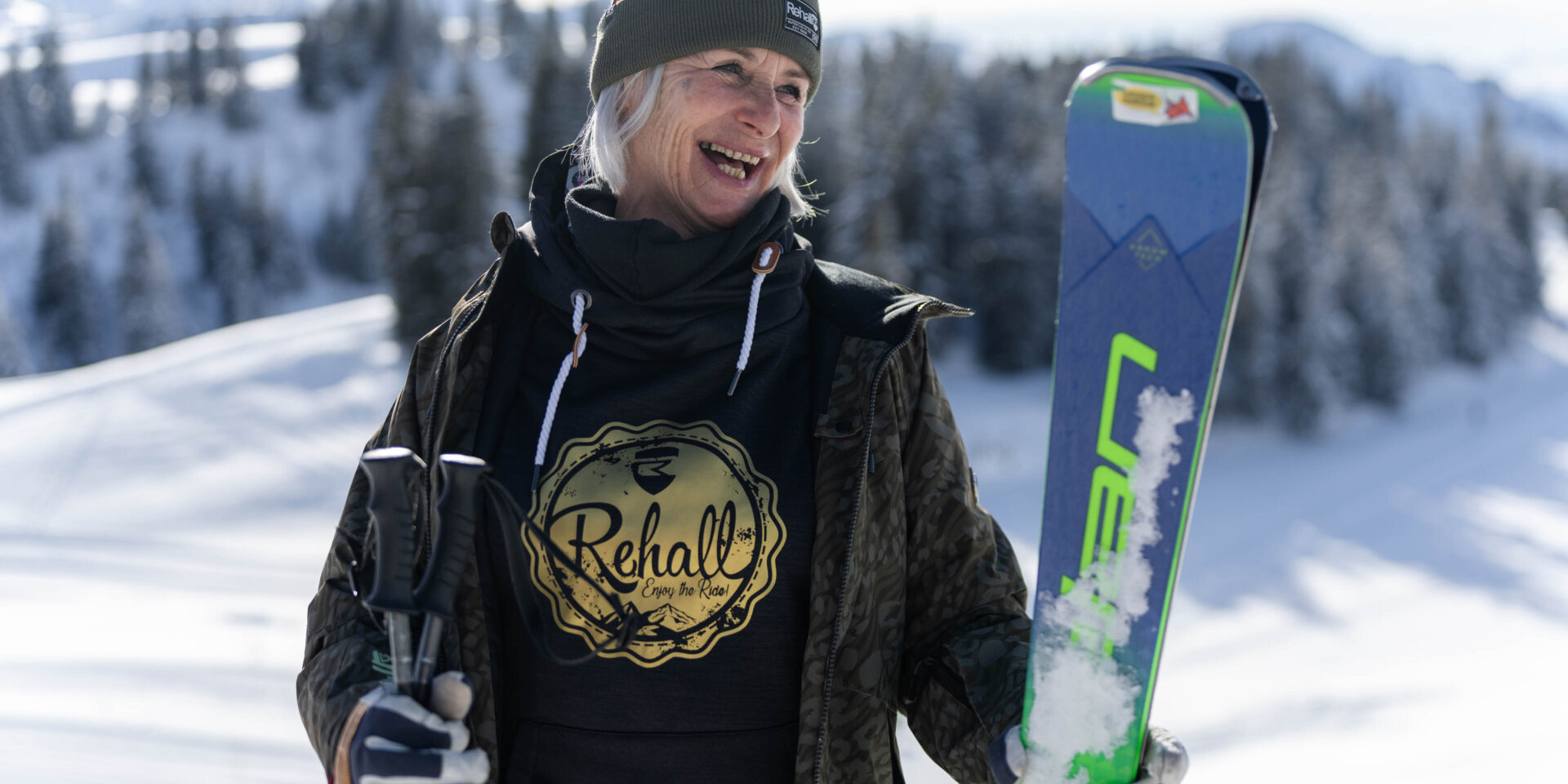 Skifahrerin steht mit offener Jacke in je einer Hand Ski und Stoecke schaut auf dei Seite und lacht