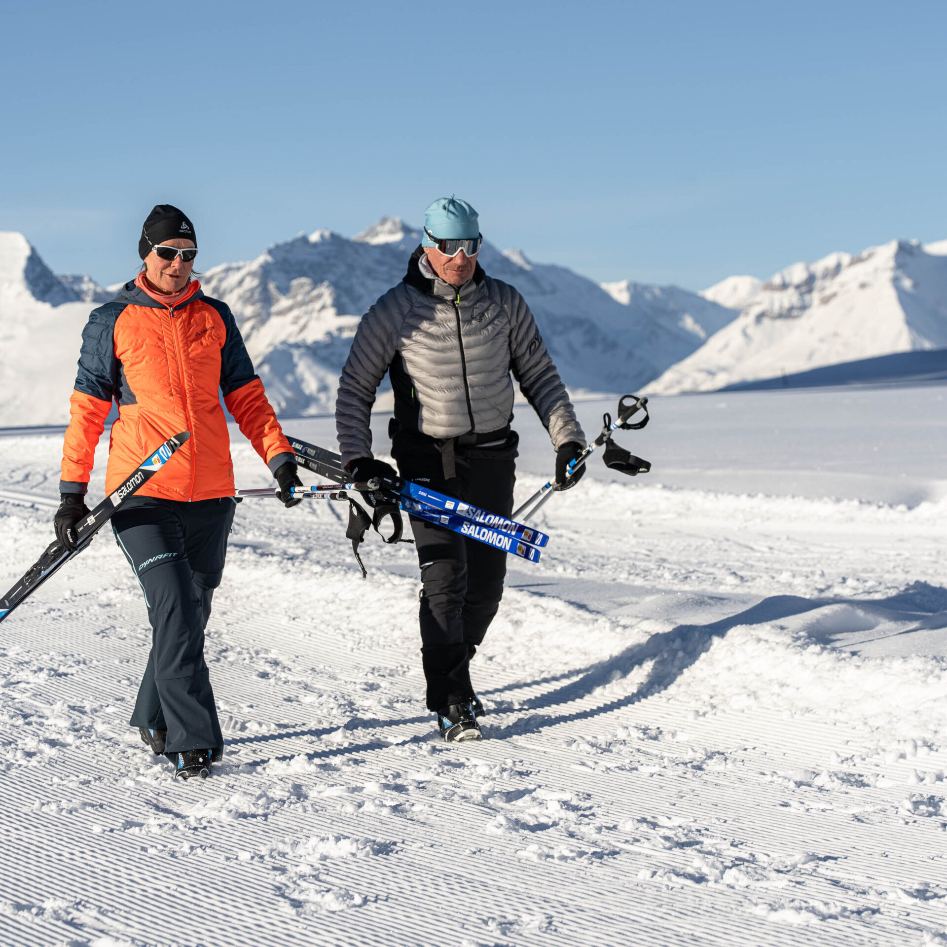 Zwei Langlaeufer tragen ihre Skis und laufen vor dem Panorama